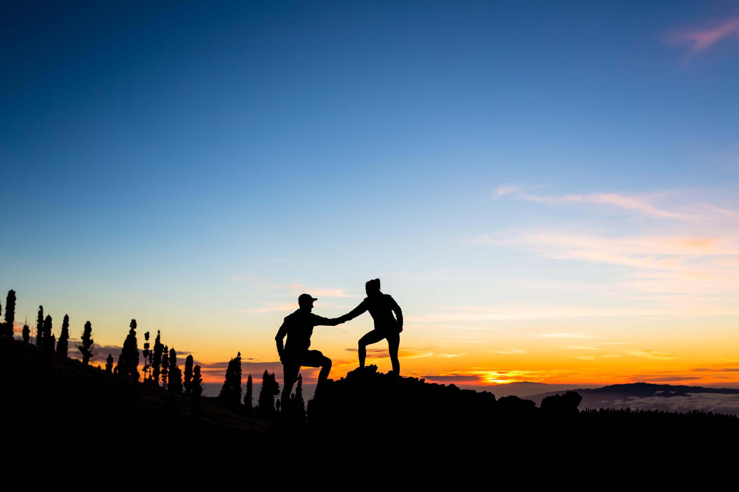 Die Schattenrisse von zwei Bergsteigenden, die sich die Hand geben vor einem Sonnenuntergang.