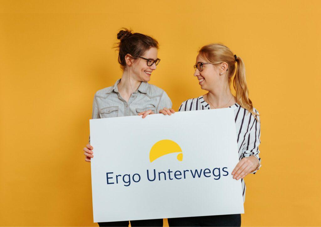 Sarah und Sara lachen sich an und halten ein großes Schild auf dem das Ergo Unterwegs Logo abgebildet ist.